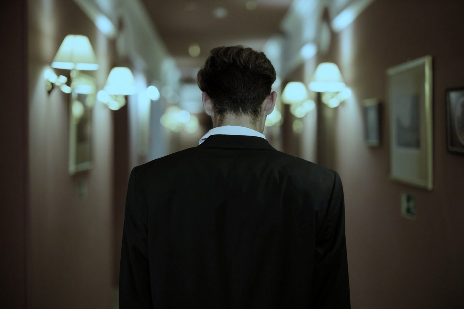 A figure seen from behind, walking a long lamplit corridor